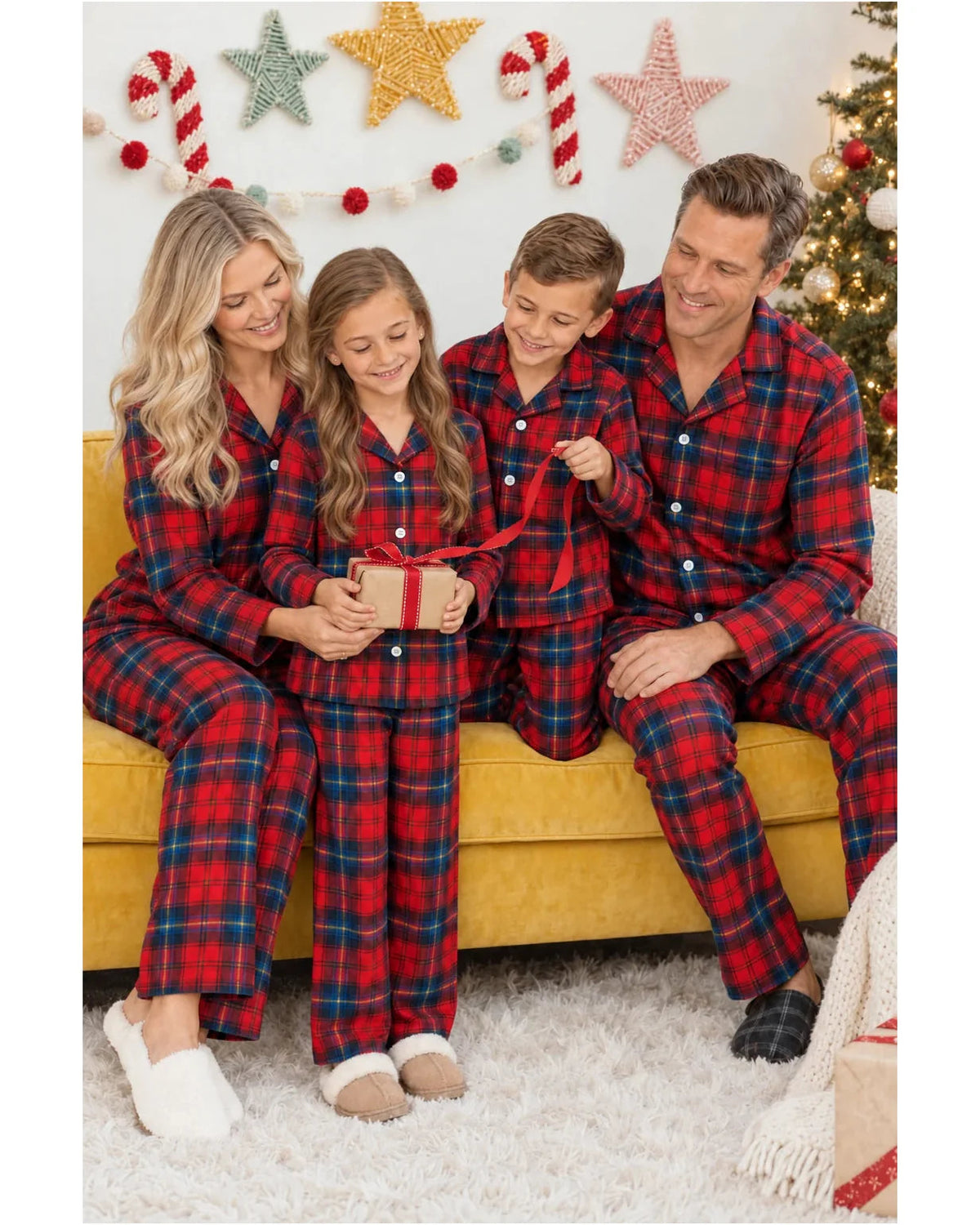 Parents and children in red and blue plaid pajamas opening a gift on a yellow sofa.