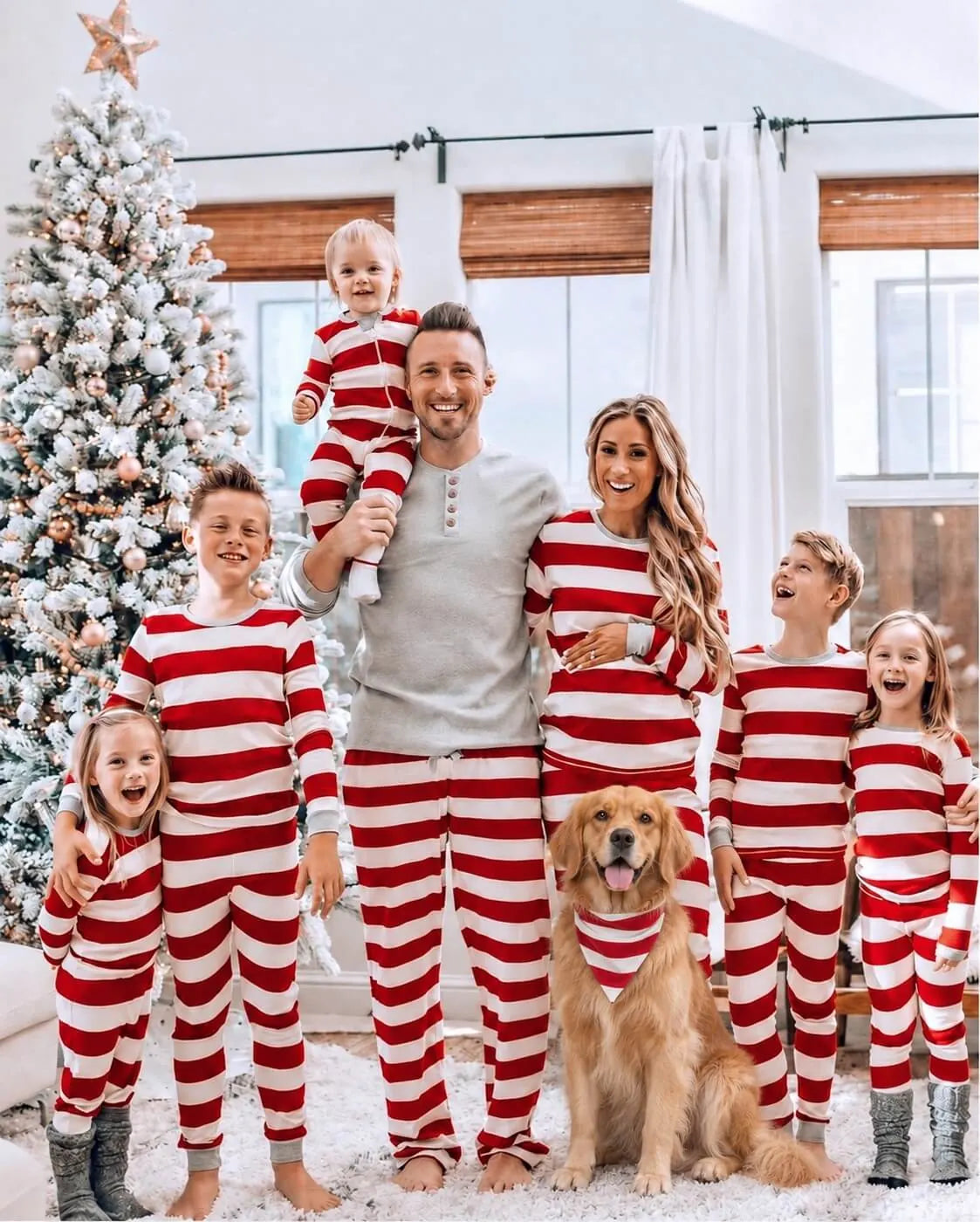 Family in red and white striped pajamas with a gray top on the man, and a dog in a striped bandana by a tree.