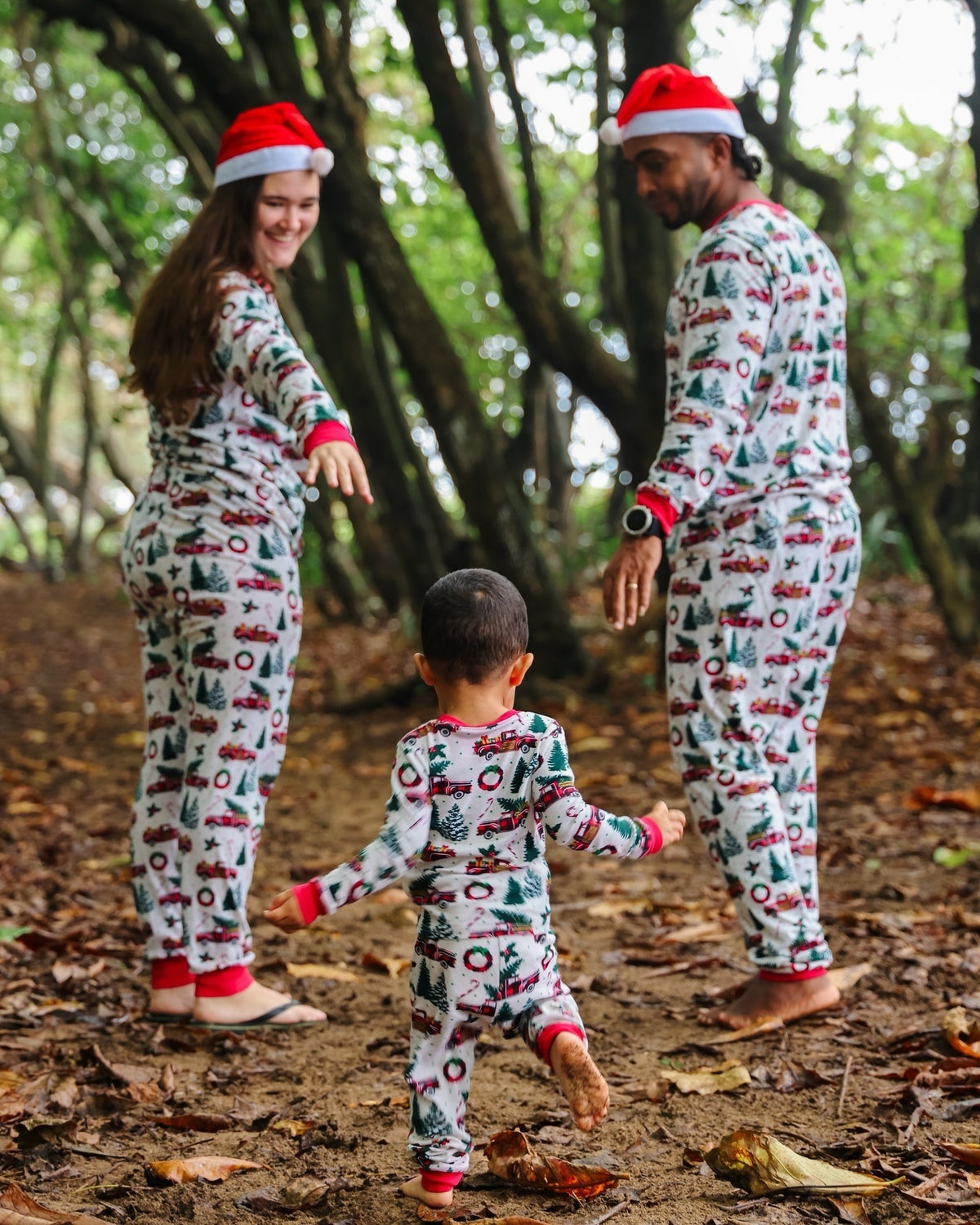 Parents reaching toward a toddler on a wooded path in white Christmas pajamas with tree and wreath prints and red cuffs.