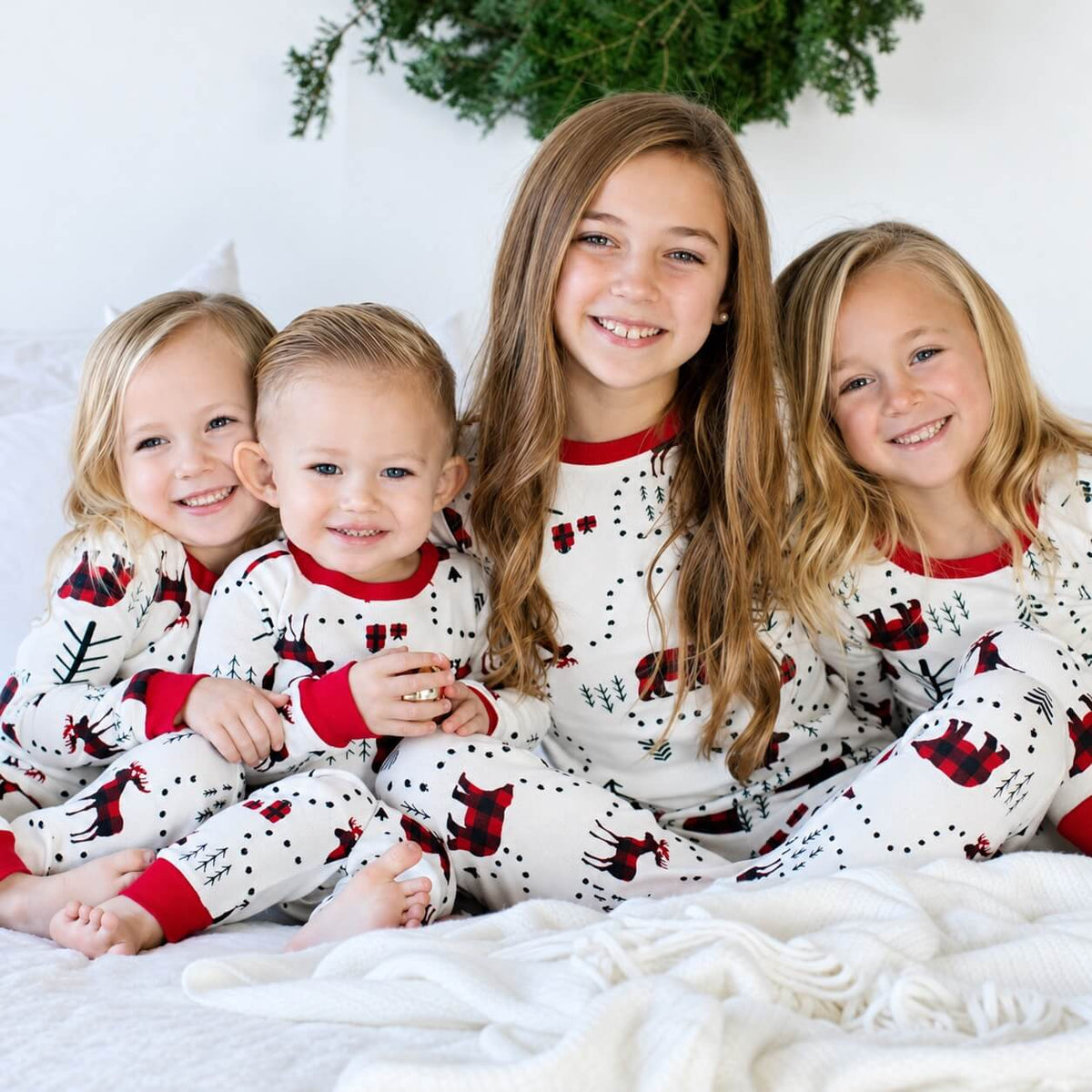 Four children sitting together in white Christmas pajamas with red cuffs and plaid animal prints.