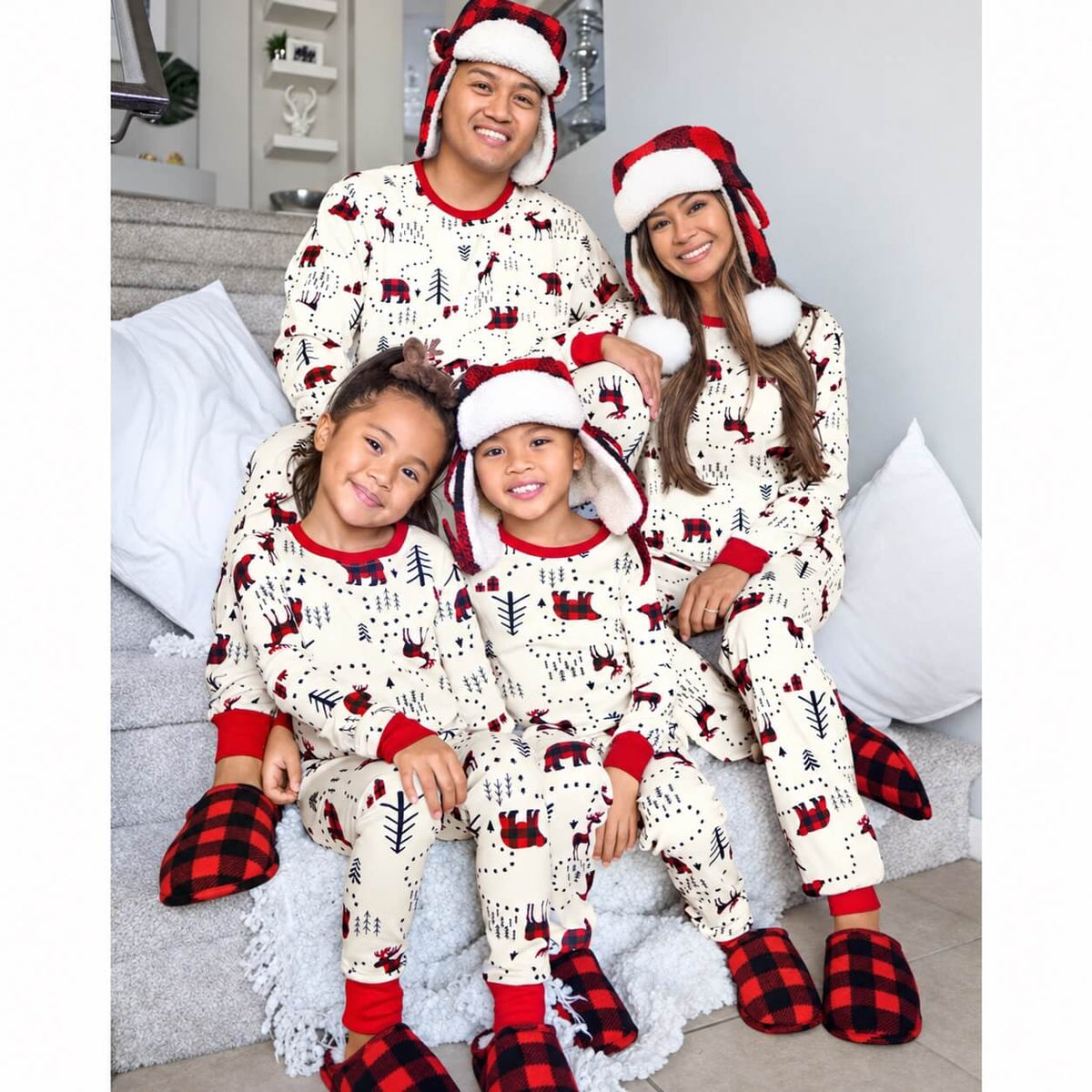 Family seated on steps in white woodland-print Christmas pajamas with red cuffs and holiday hats.