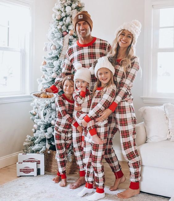 Genitori e tre bambini accanto a un albero, in pigiami natalizi a quadri beige, rossi e neri e berretti lavorati a maglia, con biscotti in mano.